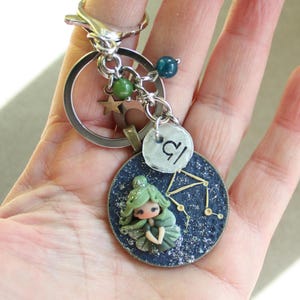 Op de afbeelding: Handgemaakte Weegschaal dierenriem sleutelhanger. Het heeft een miniatuur feeënfiguur met groen haar en jurk. De sleutelhanger bevat een zilverkleurige ring, een sterbedel en een kleine ronde bedel met het Weegschaal symbool.