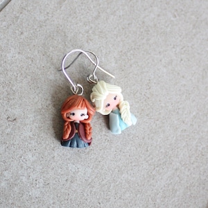 May include: A pair of dangle earrings featuring miniature figurines of two cartoon characters. One figurine has red hair and a dark outfit, while the other has blonde hair and a blue dress. The earrings have silver-colored hooks.