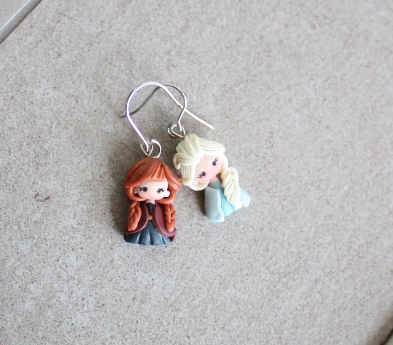 May include: A pair of dangle earrings featuring miniature figurines of two cartoon characters. One figurine has red hair and a dark outfit, while the other has blonde hair and a blue dress. The earrings have silver-colored hooks.