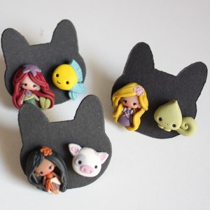 May include: Three pairs of handmade polymer clay earrings featuring Disney characters. The first pair features Ariel from The Little Mermaid with a yellow fish. The second pair features Rapunzel from Tangled with a green chameleon. The third pair features Moana from Moana with a white pig.
