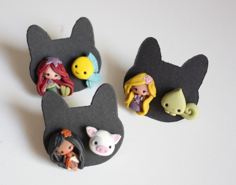 May include: Three pairs of handmade polymer clay earrings featuring Disney characters. The first pair features Ariel from The Little Mermaid with a yellow fish. The second pair features Rapunzel from Tangled with a green chameleon. The third pair features Moana from Moana with a white pig.