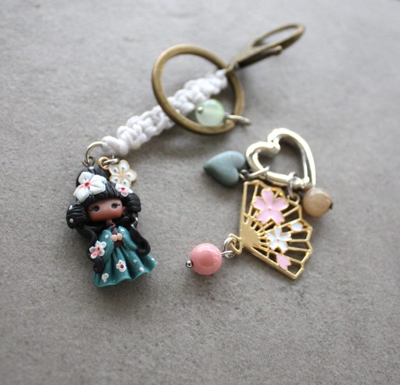 Op de afbeelding: Een sleutelhanger met twee bedels. E&eacute;n bedel is een pop in een turquoise jurk met witte bloemaccenten. De andere bedel is een gouden waaier met roze en witte bloemdetails. Een wit macram&eacute; koord verbindt de bedels met een bronzen sleutelring.