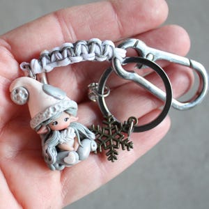 Op de afbeelding: Een handgemaakte sleutelhanger met een elfenfiguur met een roze hoed en grijs haar. De sleutelhanger heeft een zilverkleurige sluiting, een sneeuwvlokbedel en een gevlochten wit en grijs koord. De elf houdt een hart vast.