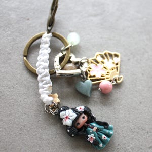 Op de afbeelding: Een decoratieve sleutelhanger met een miniatuurpop met zwart haar en een bloemenhaarstuk, gekleed in een turquoise jurk met witte bloemen. De sleutelhanger bevat een gouden waaierbedel, een hartbedel en een wit gevlochten koord.