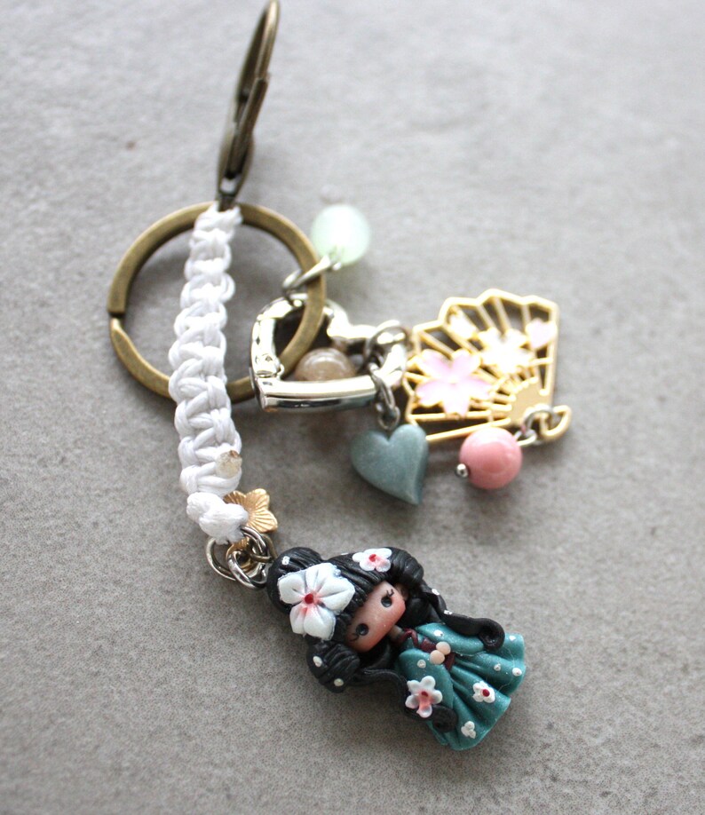 Op de afbeelding: Een decoratieve sleutelhanger met een miniatuurpop met zwart haar en een bloemenhaarstuk, gekleed in een turquoise jurk met witte bloemen. De sleutelhanger bevat een gouden waaierbedel, een hartbedel en een wit gevlochten koord.