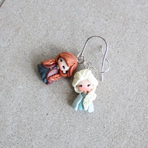 May include: A pair of handmade earrings featuring miniature doll figures. One doll has red hair in braids, the other has white hair in a braid. Both dolls wear blue and pink clothing. The earrings have silver-colored ear wires.