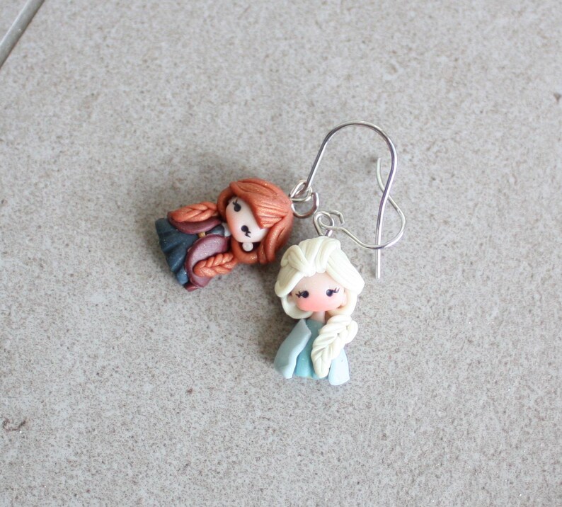 May include: A pair of handmade earrings featuring miniature doll figures. One doll has red hair in braids, the other has white hair in a braid. Both dolls wear blue and pink clothing. The earrings have silver-colored ear wires.