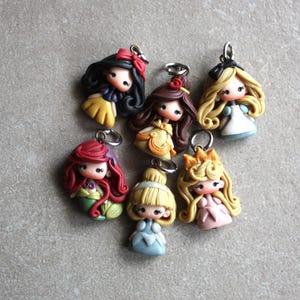 May include: Seven handmade polymer clay charms featuring Disney princesses. Each charm is a miniature figurine of a princess, including Snow White, Belle, Ariel, Cinderella, Aurora, and two others. The charms are attached to silver jump rings.