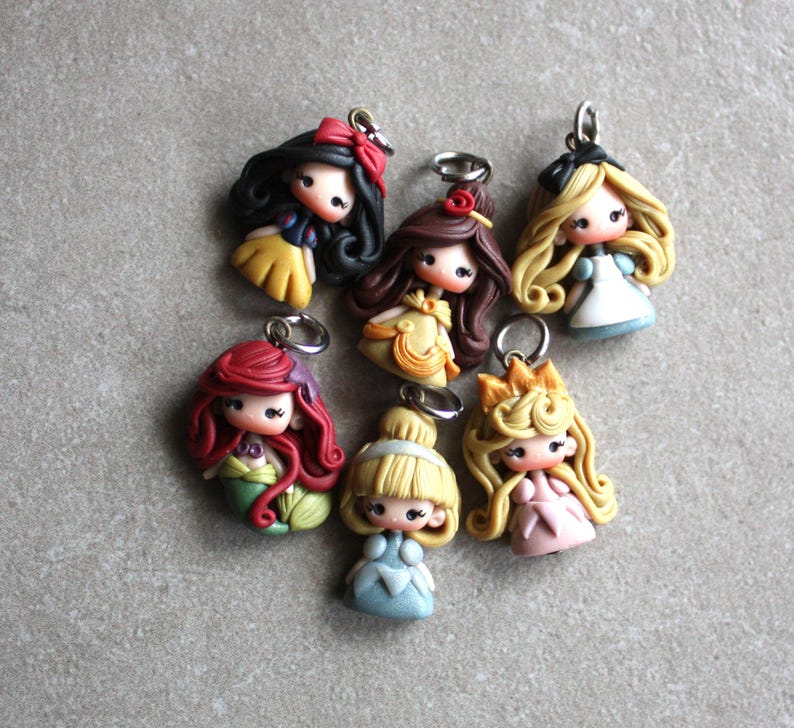 May include: Seven handmade polymer clay charms featuring Disney princesses. Each charm is a miniature figurine of a princess, including Snow White, Belle, Ariel, Cinderella, Aurora, and two others. The charms are attached to silver jump rings.