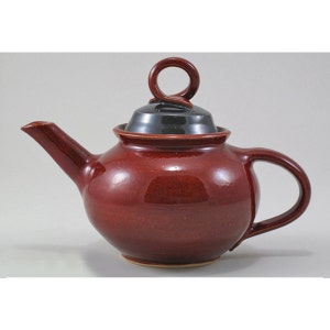 May include: A red ceramic teapot with a black lid and a looped handle. The teapot has a rounded body and a spout.