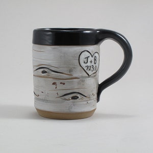May include: White ceramic mug with a black handle and a black rim. The mug has a birch bark design with a heart-shaped inscription that reads "J+B 7.23.11".