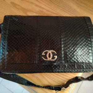 May include: A black rectangular shoulder bag with a textured snakeskin-like pattern and a glossy finish. The front flap features a prominent metallic gold-toned interlocking logo emblem in the centre. It includes a matching black shoulder strap.