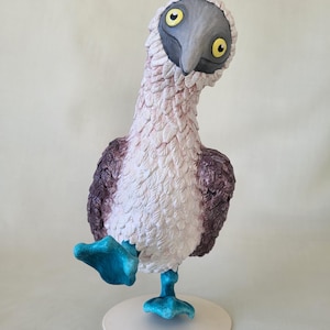 May include: A whimsical sculpture of a blue-footed booby bird. The bird has a gray head, yellow eyes, and a white and purple body. Its bright blue feet are raised in a playful pose, standing on a white circular base.
