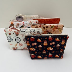 May include: A collection of zippered pouches with various fall-themed patterns. Designs include pumpkins, autumn leaves, bicycles, and coffee cups. Each pouch has a brown zipper pull and a cream-colored zipper.