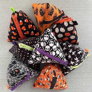May include: A set of nine Halloween-themed zippered pouches in various patterns, including skulls, candy corn, pumpkins, and ghosts. The pouches are made of fabric and have a triangular shape.