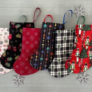 May include: Six Christmas stockings with different patterns. The stockings are made of fabric and have a loop at the top for hanging. The patterns include snowflakes, mittens, ornaments, plaid, and nutcrackers.