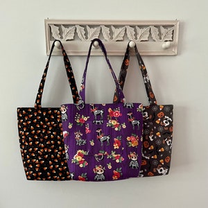 May include: Three Halloween-themed tote bags hanging on a white rack. The bag on the left has a black and orange candy corn pattern. The middle bag has a purple background with a floral pattern and skeletons. The bag on the right has a black and white spiderweb pattern with orange and white candy corn.