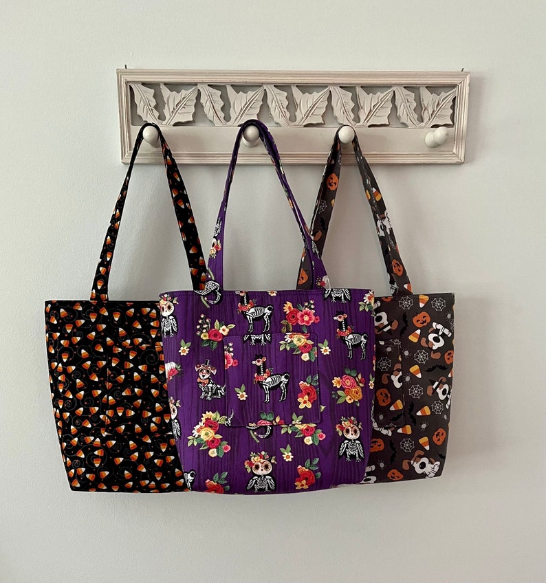 May include: Three Halloween-themed tote bags hanging on a white rack. The bag on the left has a black and orange candy corn pattern. The middle bag has a purple background with a floral pattern and skeletons. The bag on the right has a black and white spiderweb pattern with orange and white candy corn.