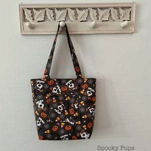 May include: A black tote bag with a Halloween-themed print featuring cartoon dogs, pumpkins, candy corn, and spiderwebs. The bag has a front pocket and a white handle.