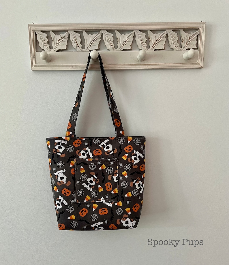 May include: A black tote bag with a Halloween-themed print featuring cartoon dogs, pumpkins, candy corn, and spiderwebs. The bag has a front pocket and a white handle.