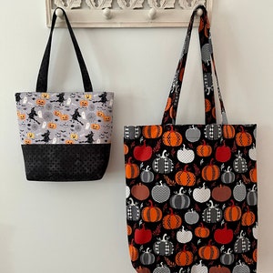 May include: Two Halloween tote bags hanging on a white wall. One bag has a gray top with witches, pumpkins, and ghosts, and a black bottom. The other bag has a black background with orange, white, and grey pumpkins.