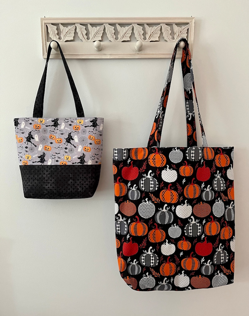 May include: Two Halloween tote bags hanging on a white wall. One bag has a gray top with witches, pumpkins, and ghosts, and a black bottom. The other bag has a black background with orange, white, and grey pumpkins.