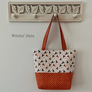 May include: A fabric tote bag with a white background, featuring a pattern of witch hats, stars, and moons. The bottom section is a solid orange with a polka dot pattern. The bag has orange handles and the text "Witchs' Hats".