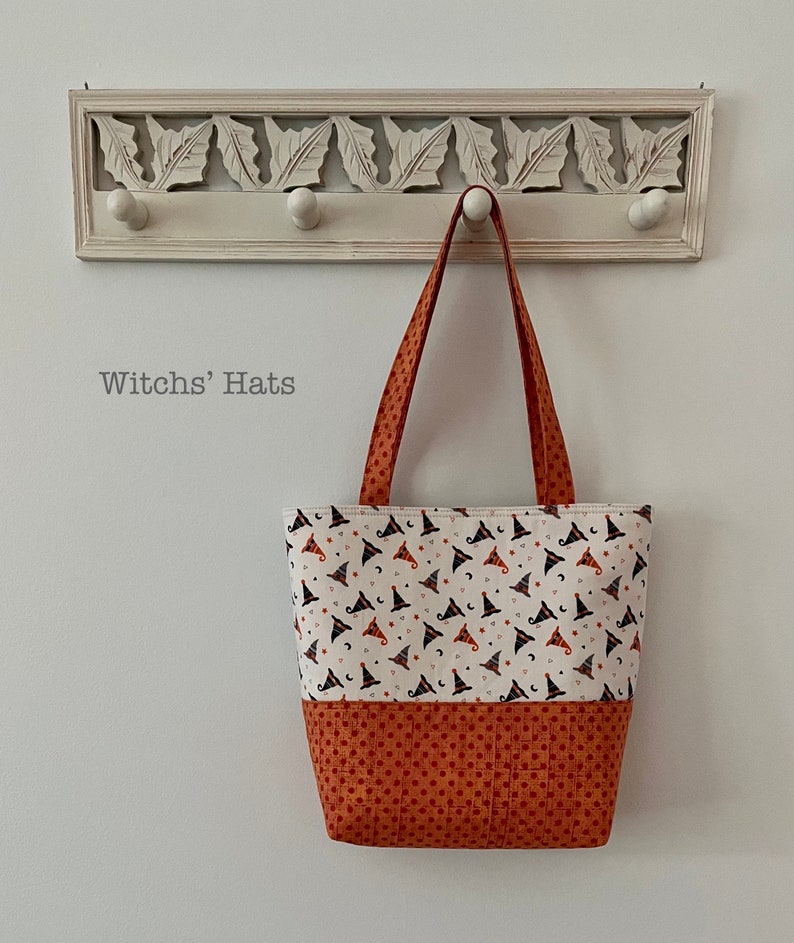 May include: A fabric tote bag with a white background, featuring a pattern of witch hats, stars, and moons. The bottom section is a solid orange with a polka dot pattern. The bag has orange handles and the text "Witchs' Hats".