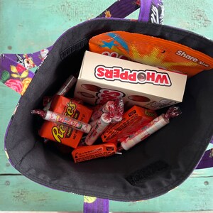 May include: A black fabric tote bag filled with various sweets, including Whoppers, Reese's Peanut Butter Cups, Smarties, and other assorted sweets. The bag has a purple and black floral print with a skull design.