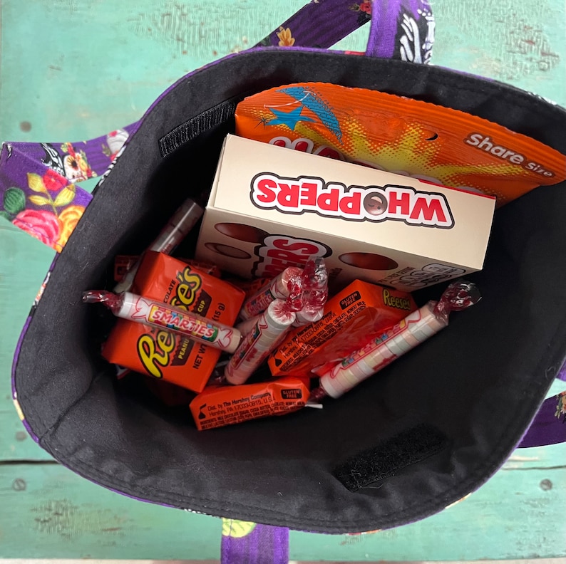 May include: A black fabric tote bag filled with various sweets, including Whoppers, Reese's Peanut Butter Cups, Smarties, and other assorted sweets. The bag has a purple and black floral print with a skull design.