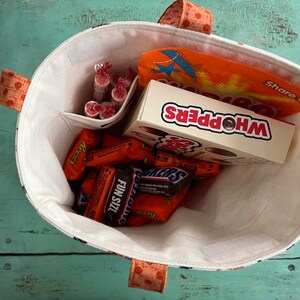 May include: A fabric tote bag with orange polka dot handles and trim, filled with various sweets. The bag contains a box of Whoppers, Snickers, Reese's, and other wrapped treats. The interior is white with a small pocket.