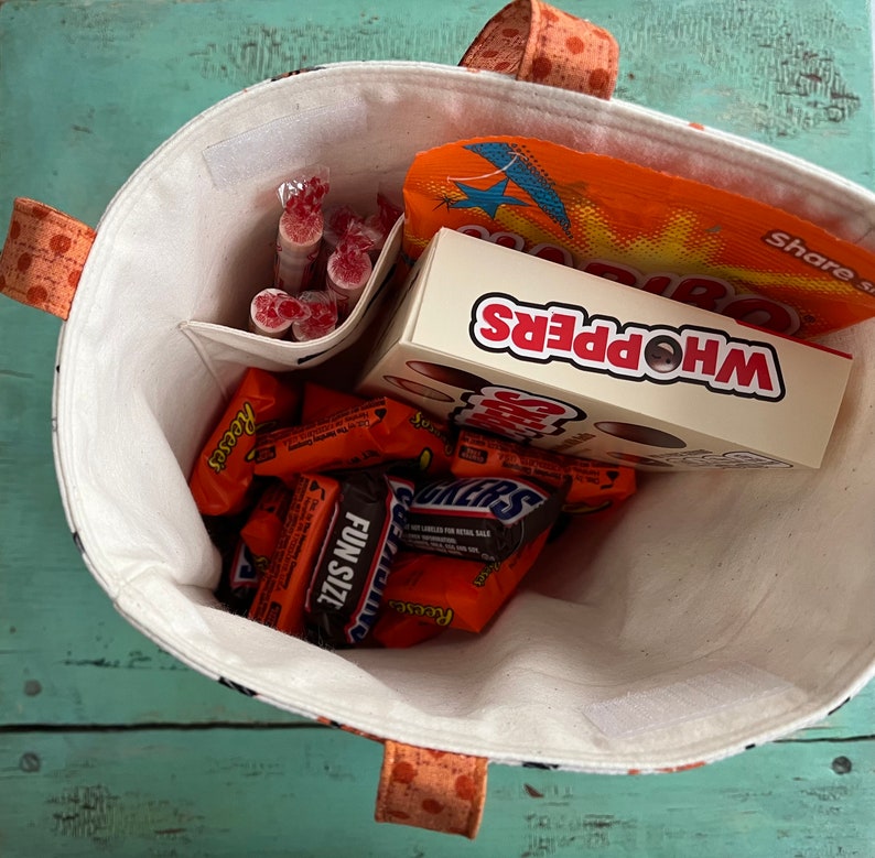 May include: A fabric tote bag with orange polka dot handles and trim, filled with various sweets. The bag contains a box of Whoppers, Snickers, Reese's, and other wrapped treats. The interior is white with a small pocket.