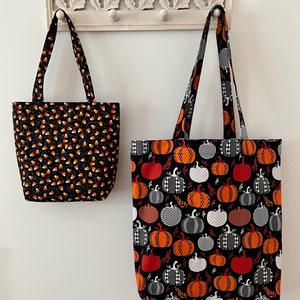 May include: Two Halloween-themed tote bags hanging on a white rack. The smaller bag is black with orange and white candy corn designs. The larger bag is black with orange, white, and grey pumpkins.