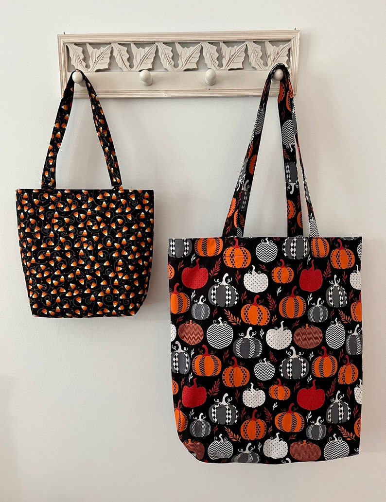 May include: Two Halloween-themed tote bags hanging on a white rack. The smaller bag is black with orange and white candy corn designs. The larger bag is black with orange, white, and grey pumpkins.