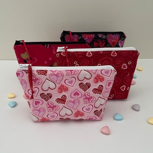 May include: Four zippered pouches with different heart patterns. The pouches are made of fabric and have a red, pink, and black color scheme.