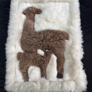 May include: A rectangular wall hanging made of white sheepskin featuring a brown, fuzzy applique of an adult llama and a smaller baby llama. The white fur creates a thick, textured border around the central brown animal figures.