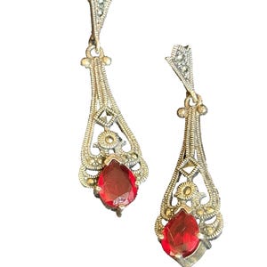 May include: A pair of vintage drop earrings. Each earring features a silver-toned filigree design with a teardrop-shaped, red gemstone. The earrings have a decorative top with small, embedded stones and a delicate, openwork pattern.