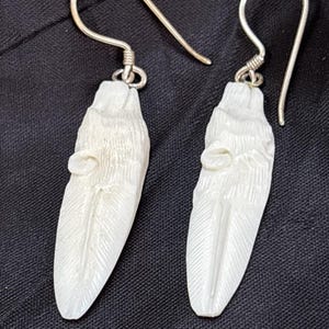 May include: A pair of drop earrings featuring carved off-white feather-shaped pendants suspended from silver-toned metal ear wires. The pendants display detailed linear textures resembling the barbs and central shaft of a bird feather.