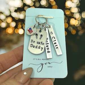 Peut inclure: Un porte-clés argenté avec une variété de breloques, dont un petit drapeau américain, un breloque avec des empreintes de pas, un breloque qui dit "Be safe Daddy", un breloque qui dit "Poland" et un breloque qui dit "Clair". Le porte-clés est sur une carte bleue avec le texte "It's all about you amy".