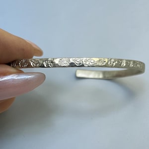May include: A silver-colored cuff bracelet featuring a hammered texture and a repeating pattern of small, embossed floral motifs along the outer surface. The bracelet has an open-ended design with rounded, smooth ends and a metallic finish.