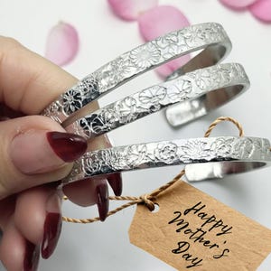 May include: Three silver-colored floral engraved bangles. Each bangle features a different floral pattern. A small brown tag with the words "Happy Mother's Day" is attached to one of the bangles with a twine string. Pink flower petals are in the background.