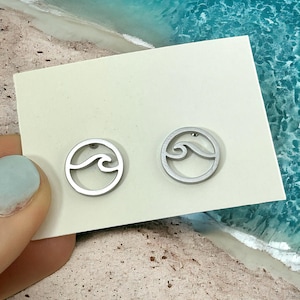 May include: A pair of silver earrings with a wave design. The earrings are on a white card against a background of sand and blue water.