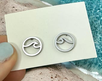 May include: A pair of silver earrings with a wave design. The earrings are on a white card against a background of sand and blue water.