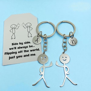 May include: Two silver keychains with stick figure charms. Each keychain has a round charm with a letter, "A" and "n". A card with stick figures and the text "Side by side, we'll always be... Flipping off the world, just you and me." is in the background.