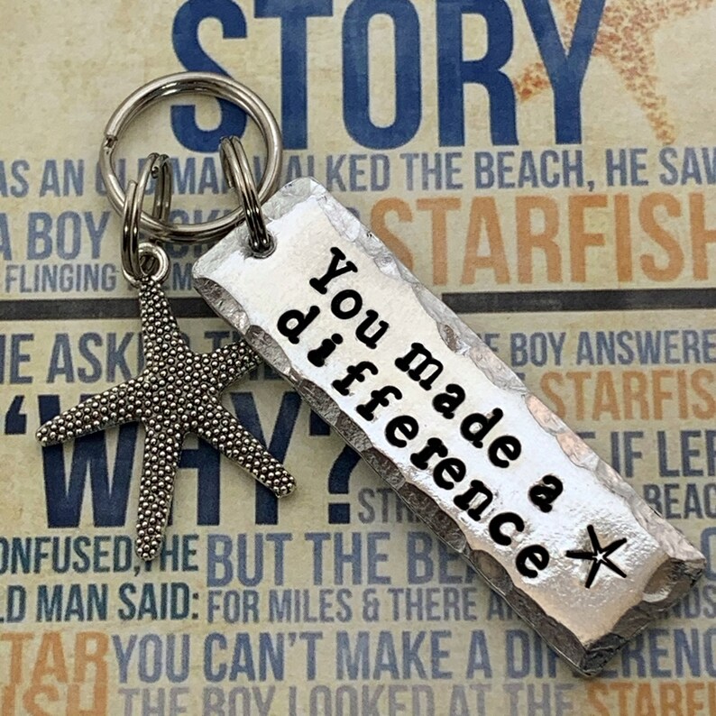 May include: A silver-toned metal keychain featuring a rectangular pendant with hammered edges and the black engraved text You made a difference followed by a small star symbol. A separate silver-toned starfish charm hangs from the metal ring.