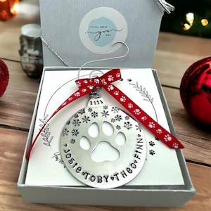 May include: A silver Christmas ornament with a paw print cutout and snowflake details, personalized with names. It's tied with a red ribbon featuring white paw prints, presented in a gift box. The ornament is approximately 3 inches in diameter.