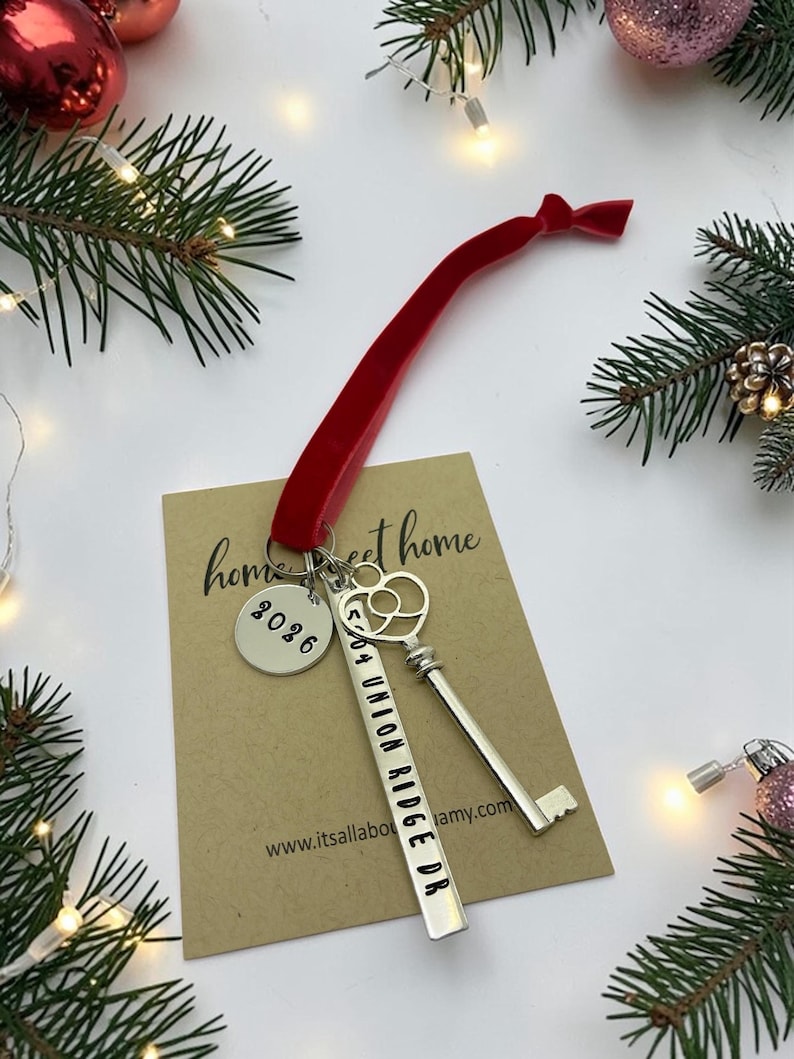 a custom new home ornament with a long silver tag with the street address hand stamped, a circle tag with the current year the home was purchased, a silver skeleton key, and a plush red velvet ribbon. set on a white background with Christmas tree evergreen branches and soft twinkling lights and red ornaments
