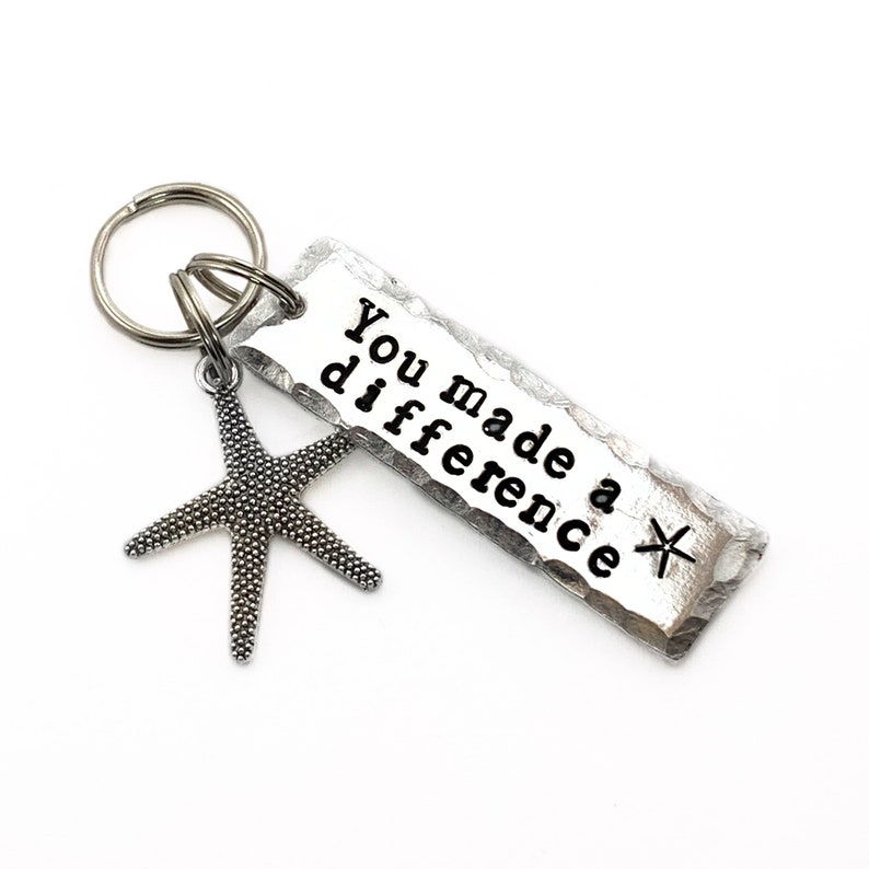 May include: A silver-toned metal keychain featuring a rectangular tag with a hammered texture and the stamped black text "You made a difference" followed by a small star symbol. A textured starfish charm is attached to the circular split ring.