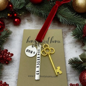 a custom new home ornament with a long silver tag with the street address hand stamped, a circle tag with the current year the home was purchased, a gold skeleton key, and a plush red velvet ribbon. set on a white background with Christmas tree evergreen branches and soft twinkling lights and red ornaments
