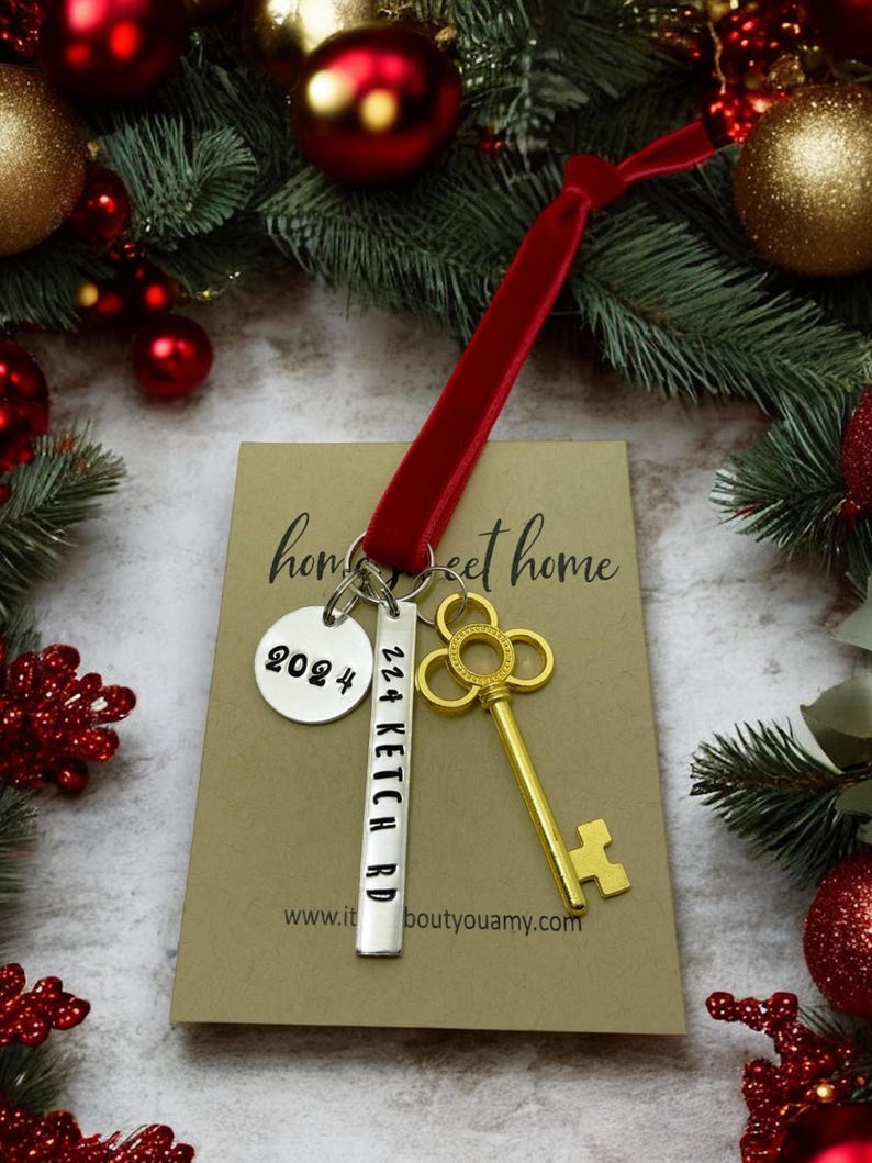 a custom new home ornament with a long silver tag with the street address hand stamped, a circle tag with the current year the home was purchased, a gold skeleton key, and a plush red velvet ribbon. set on a white background with Christmas tree evergreen branches and soft twinkling lights and red ornaments